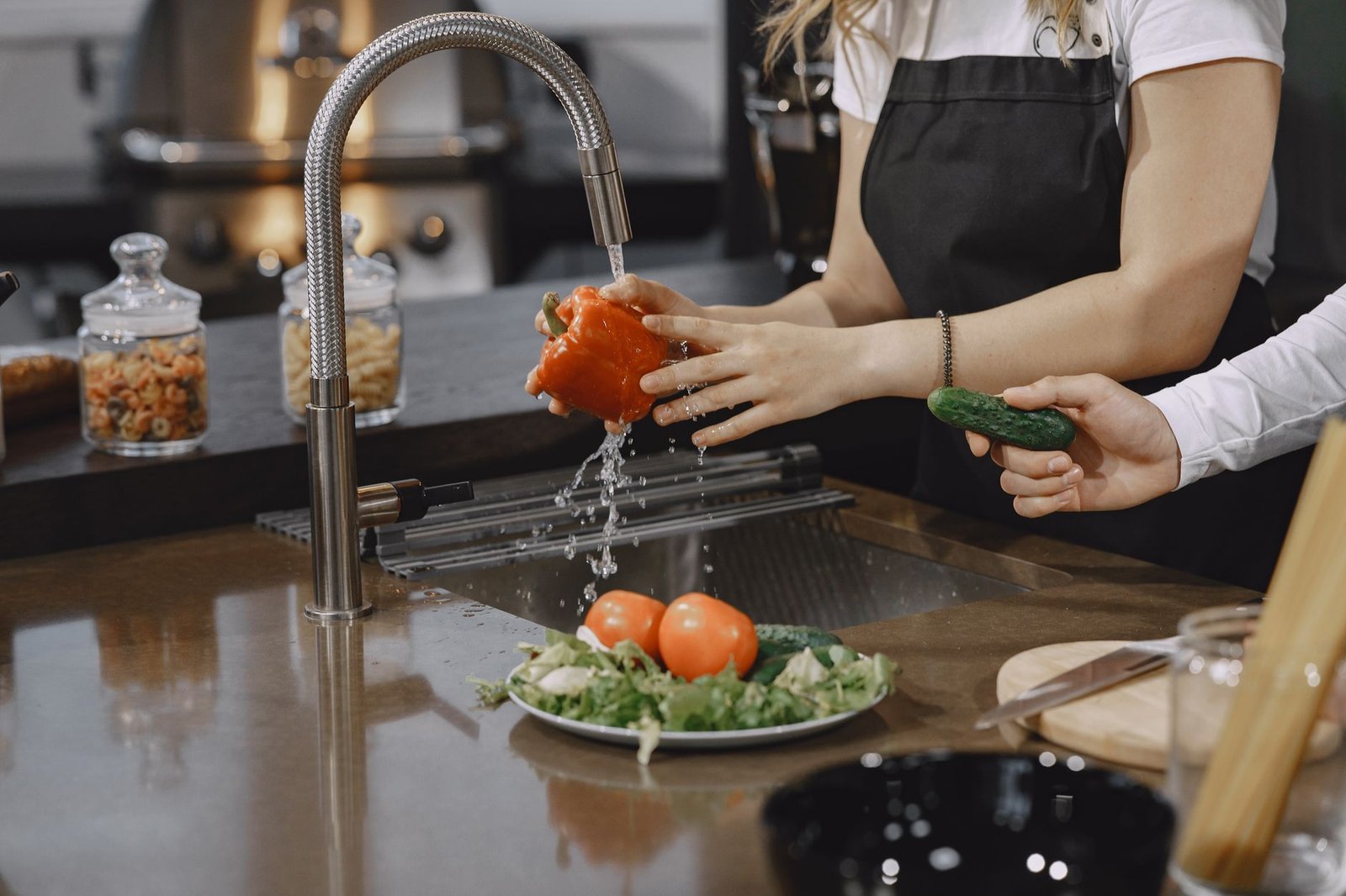 Hands washing a red bell pepper in a modern kitchen sink with fresh vegetables.