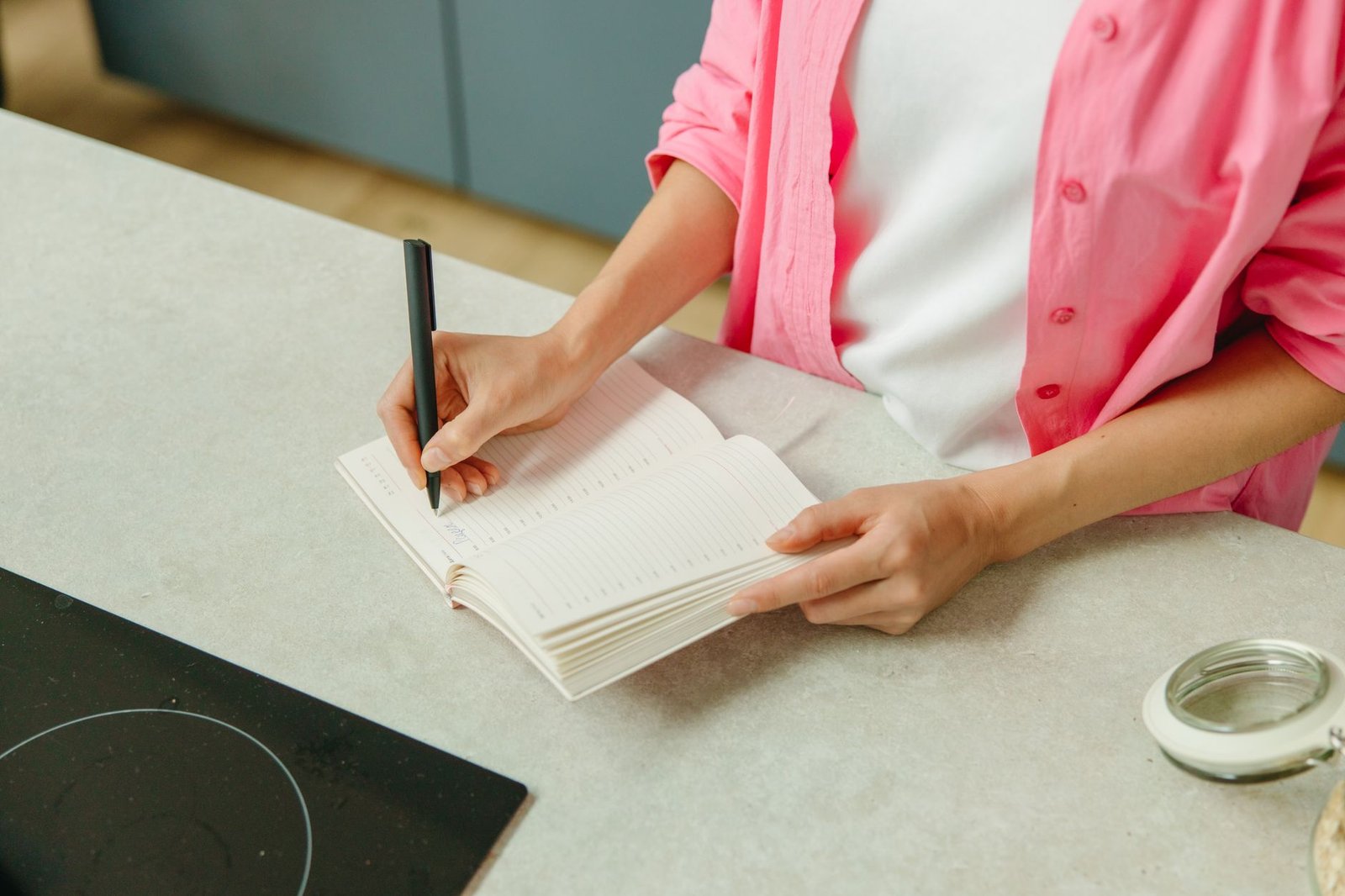A woman in a pink shirt writing in a notebook on a kitchen counter with a pen.