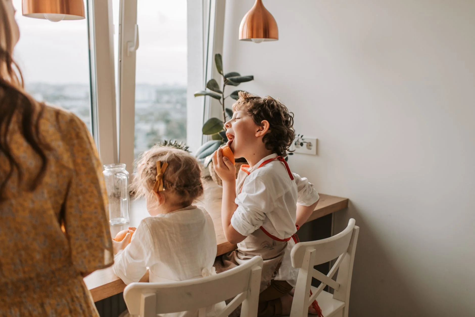 Two children enjoying an orange snack indoors by a window with natural light.