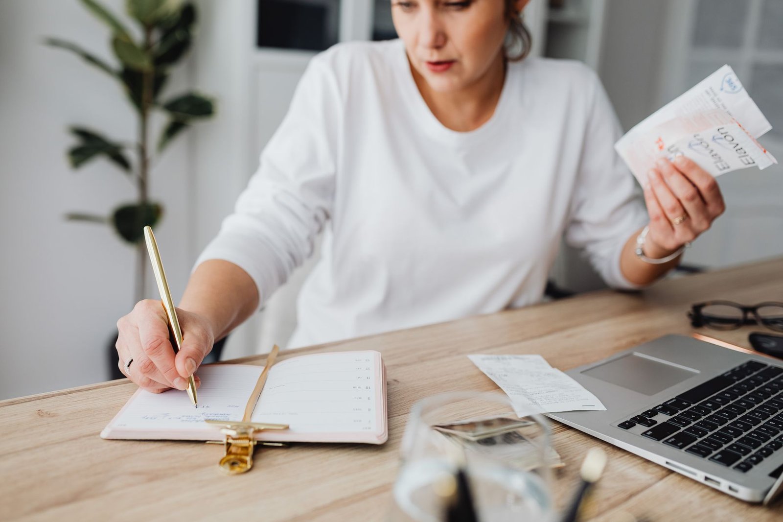 budgeting with irregular income — Woman managing finances, writing on notepad, and holding receipts at a desk.