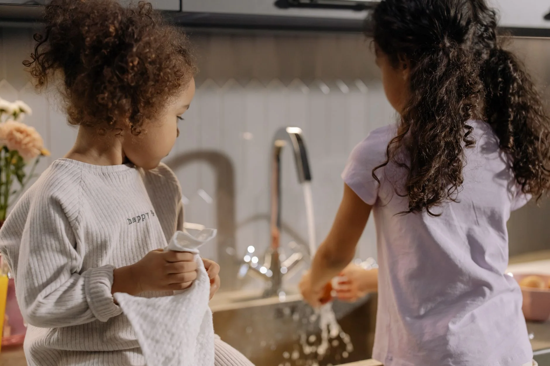 chore chart by age — Two young girls washing dishes together in a cozy kitchen setting.