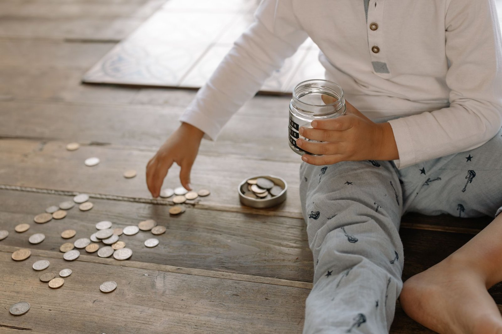 emergency fund — A young child collects coins into a jar on a wooden floor, symbolizing savings.