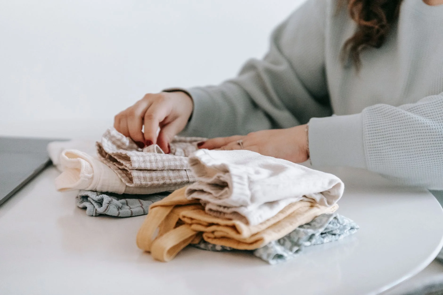 Crop faceless female in casual clothes folded colorful baby garments while sitting at white table in room on white background