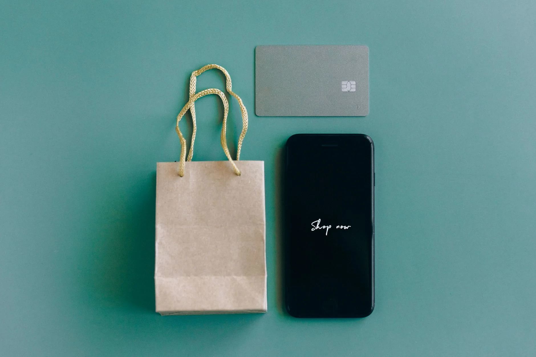 how to stop impulse spending — Top view of smartphone with 'Shop now' text, credit card, and paper bag on green background.