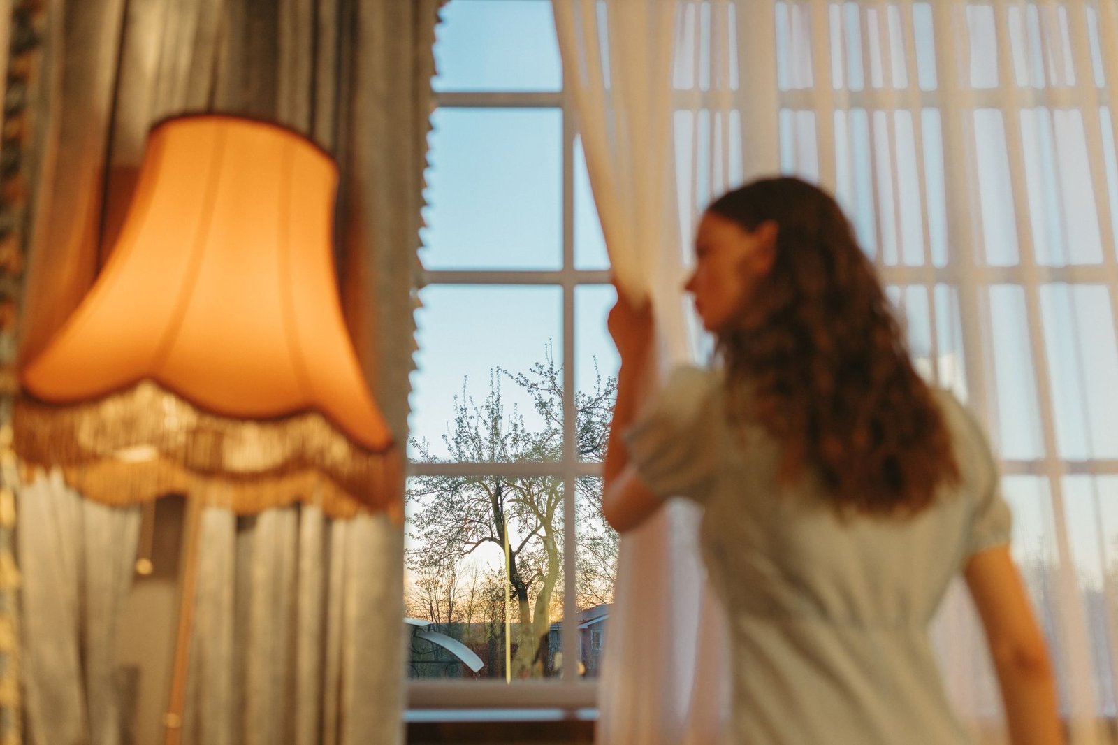 A woman looks out a window at sunset, drawing back curtains in a cozy room.