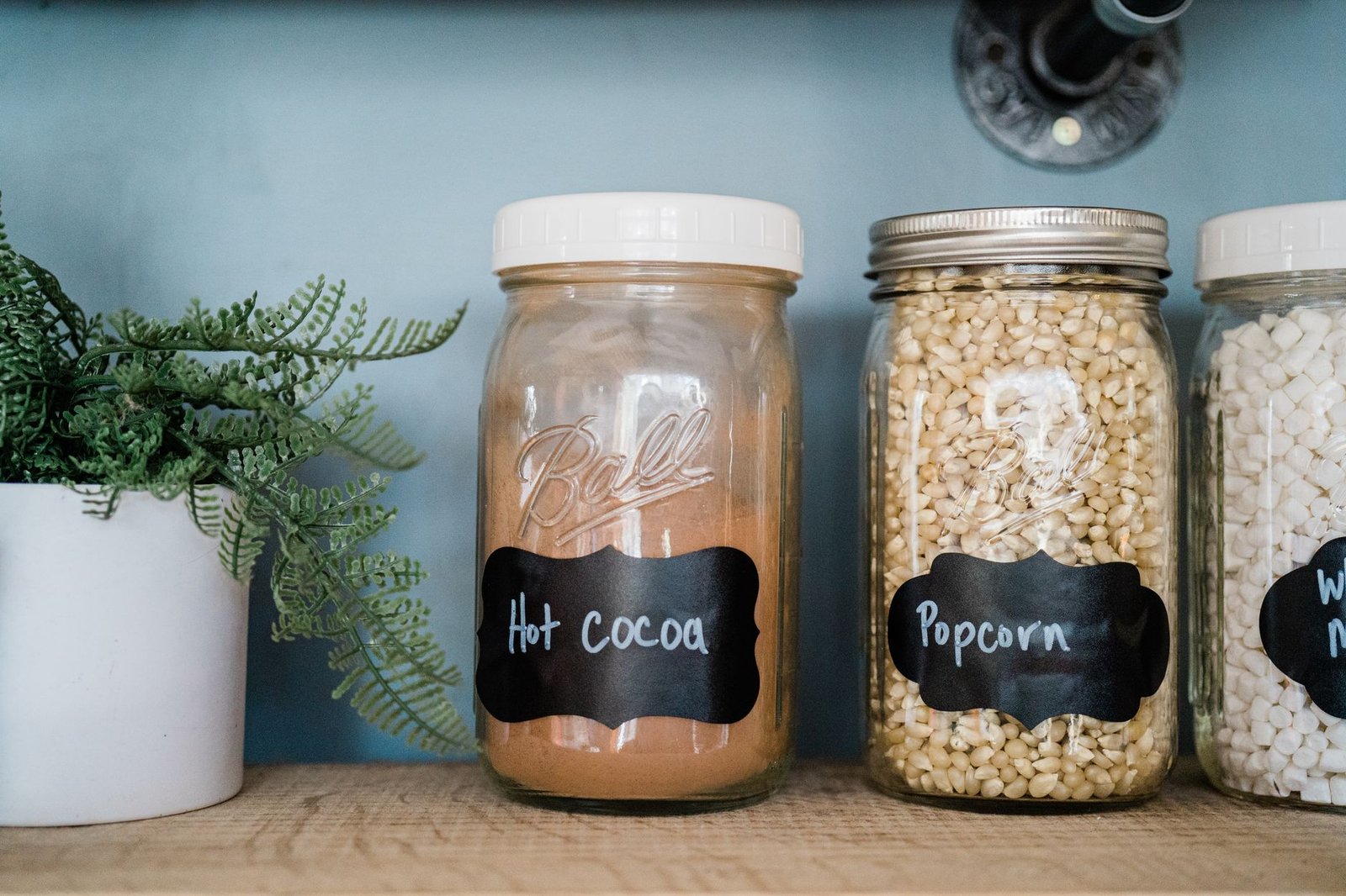 pantry staples list — Glass jars with popcorn, hot cocoa, and marshmallows on a kitchen shelf with a potted plant.