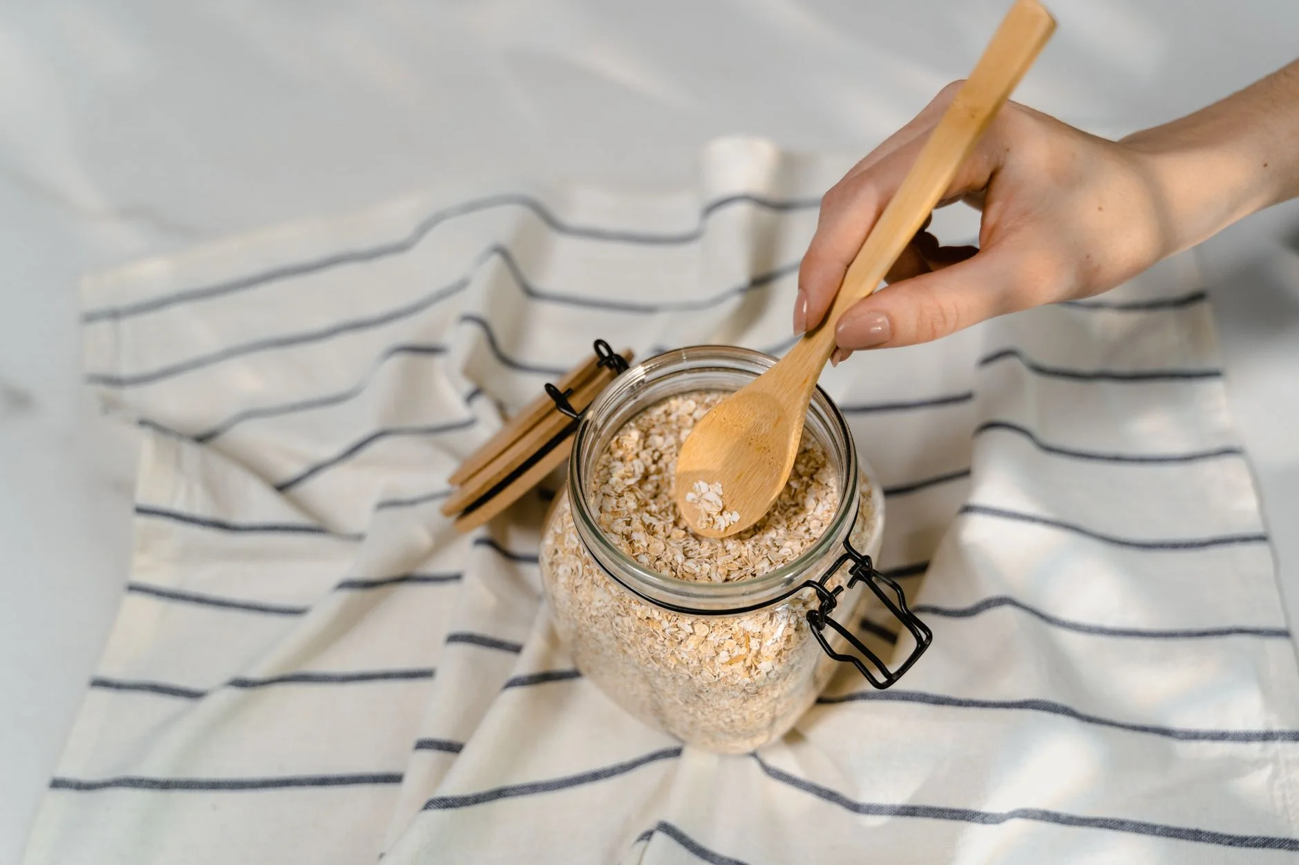 quick breakfast ideas — Close-up of a hand using a wooden spoon to scoop uncooked oatmeal from a glass jar on a striped cloth.
