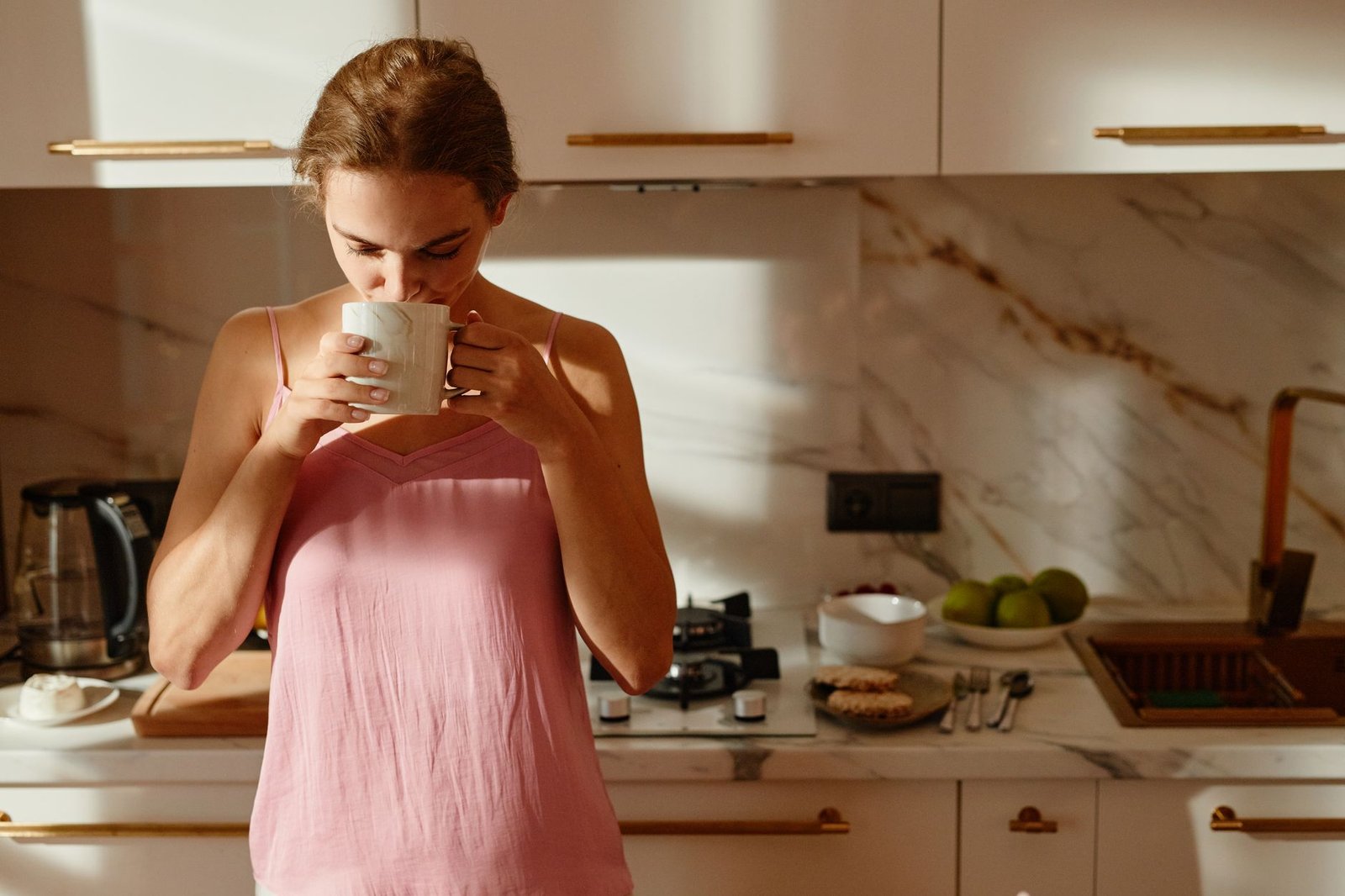 realistic morning routine — A woman enjoys a hot beverage in a sunlit kitchen, creating a tranquil morning scene.