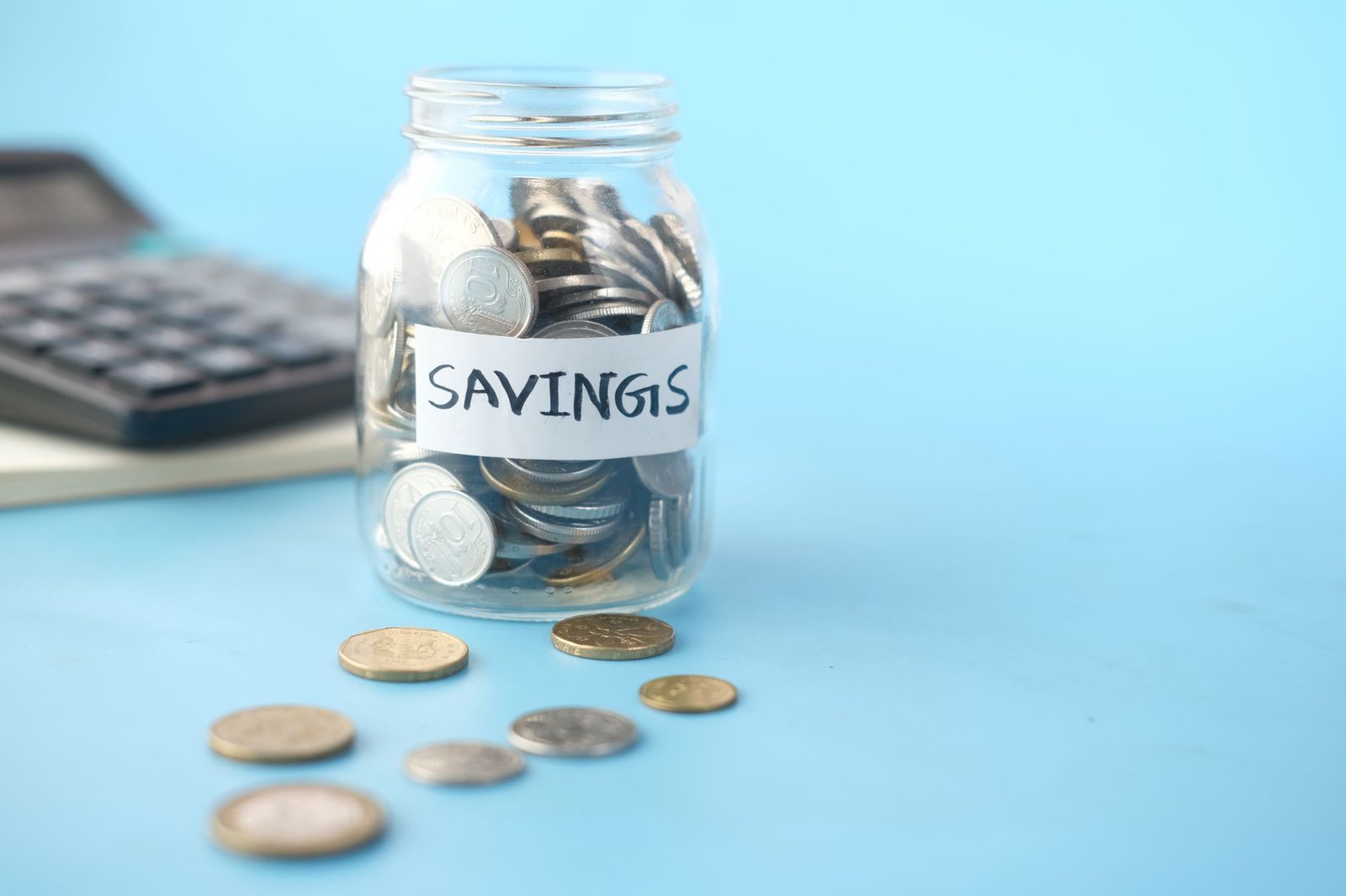 sinking funds — Glass jar labeled 'Savings' filled with coins, beside a calculator on a blue background.