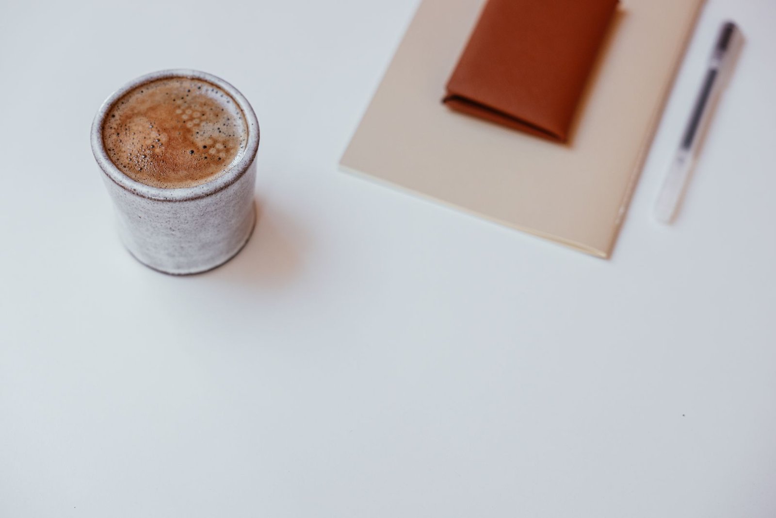A minimalist setup with a cup of coffee, a notebook, and a pen on a white tabletop.