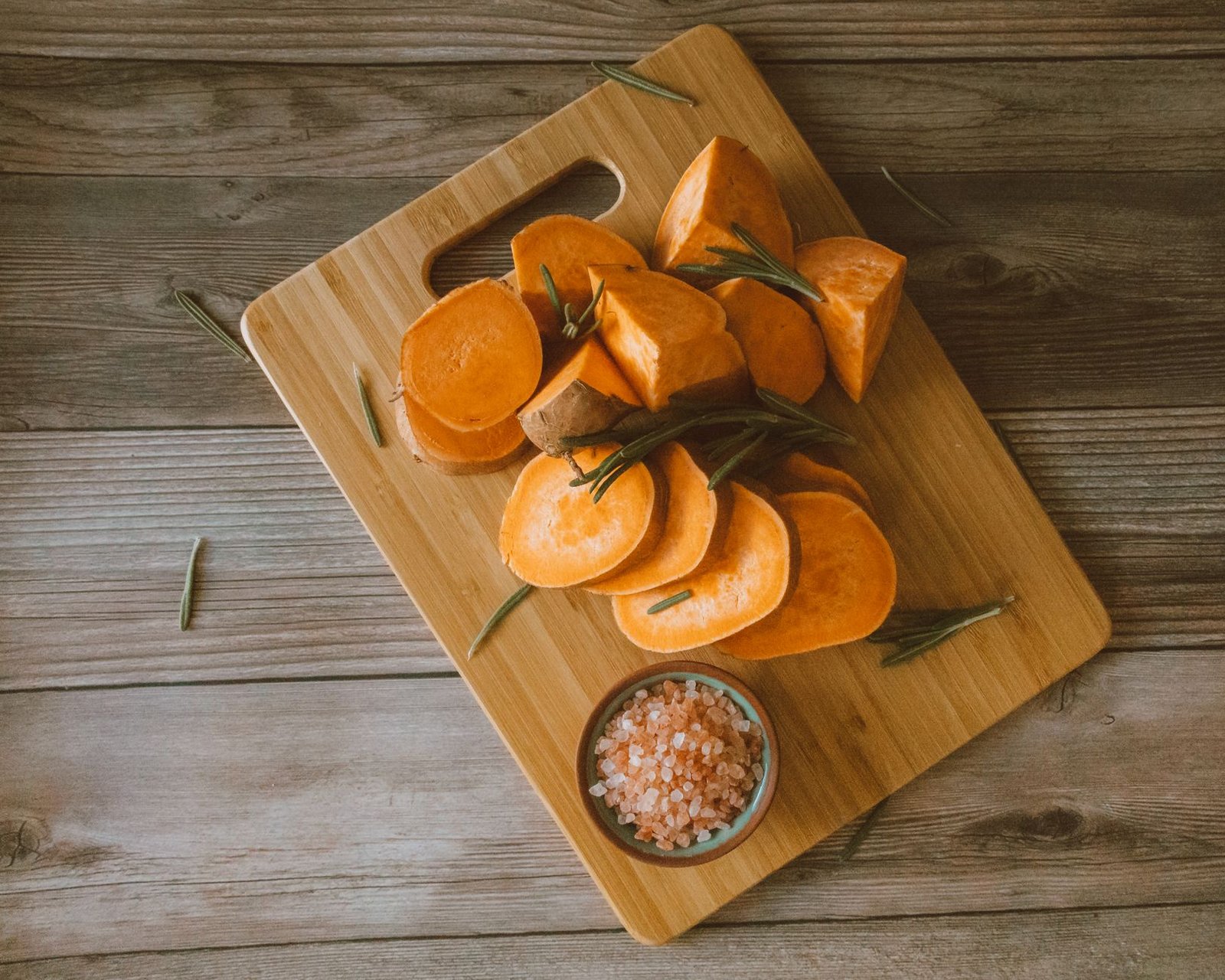 Chopped sweet potatoes on a bamboo board with Himalayan salt and rosemary, perfect for rustic food themes.