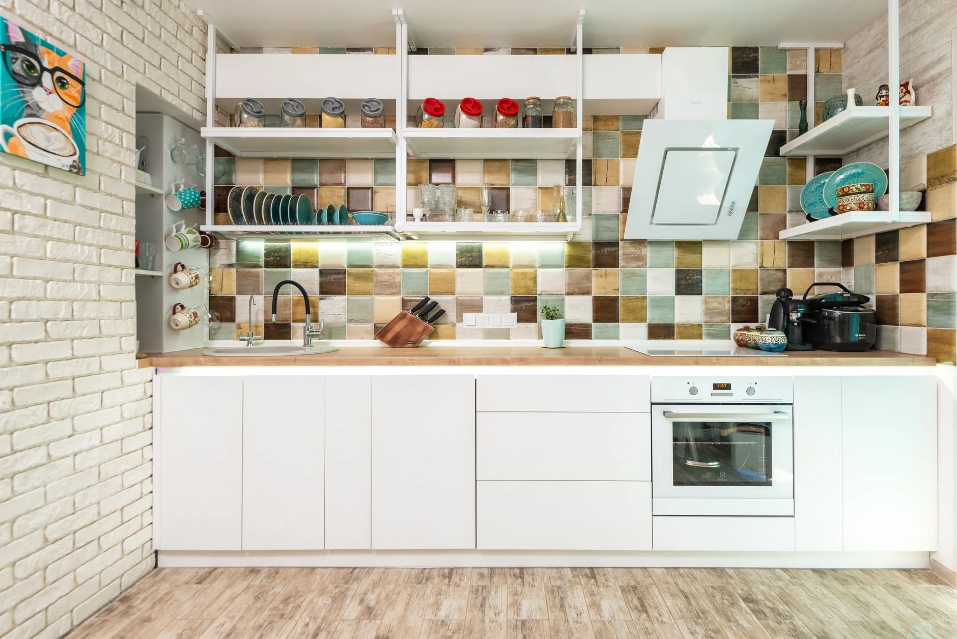 Interior of bright kitchen furnished with white cupboards and shelves with oven decorated with various dishware and utensils with tiled multicolored backsplash