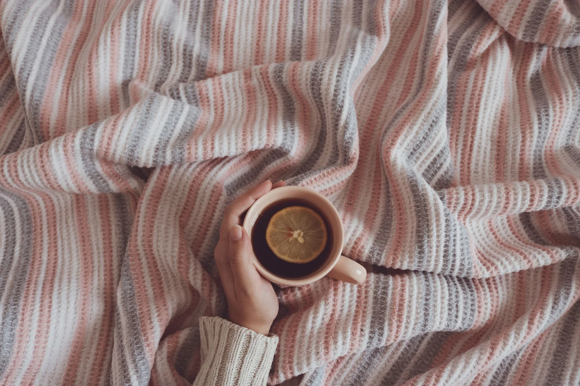 Hand holding cup of tea with lemon on a cozy striped blanket, evoking warmth and comfort.
